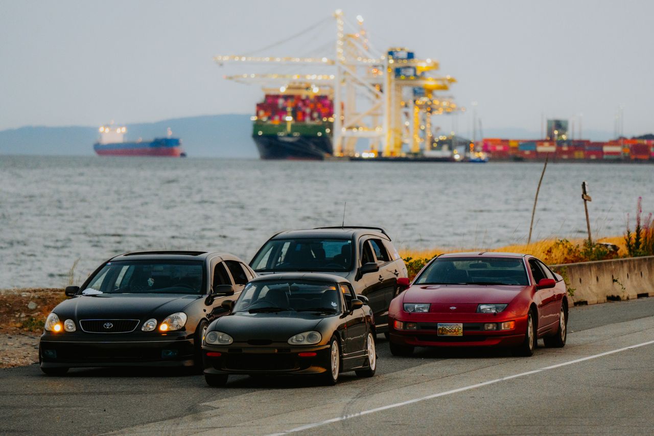 Cars lined up near a bustling port with large ships and cranes visible in the background.