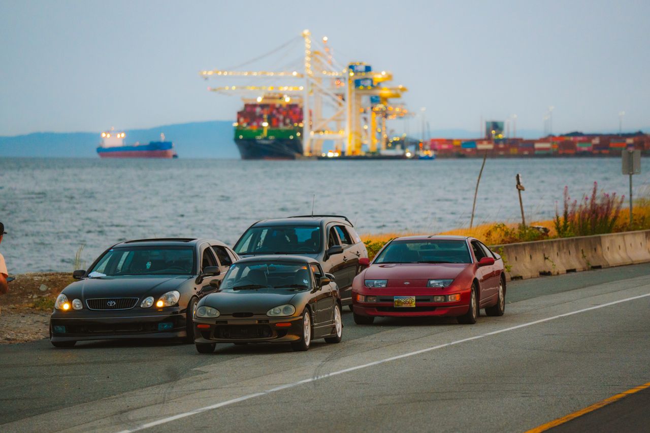 Three parked cars by the seashore with cargo ships and cranes at a harbor nearby, during the day.
