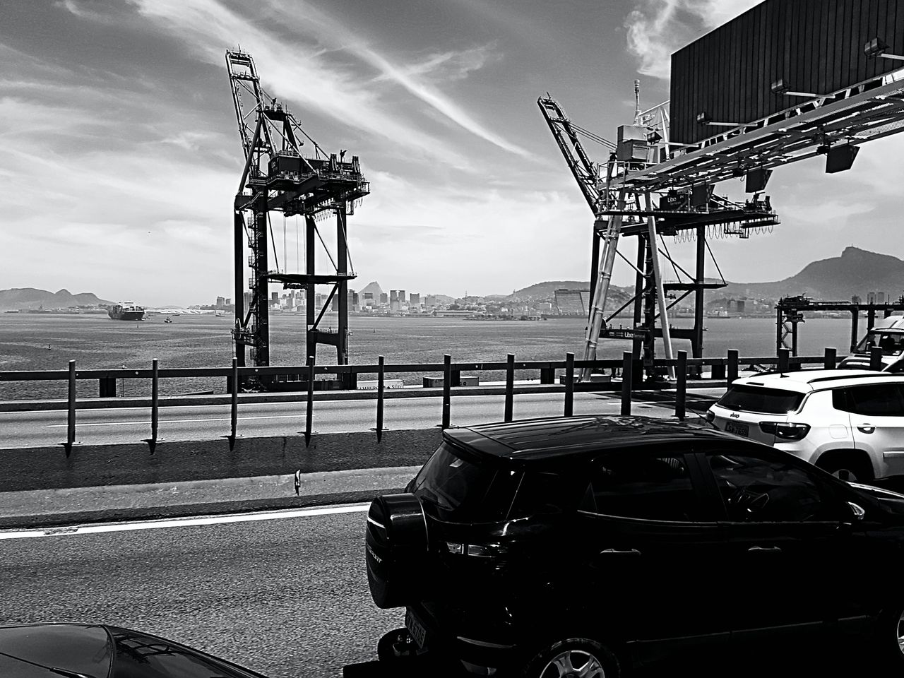 Black and white image of a bustling harbor with cranes, cars, and cityscape.